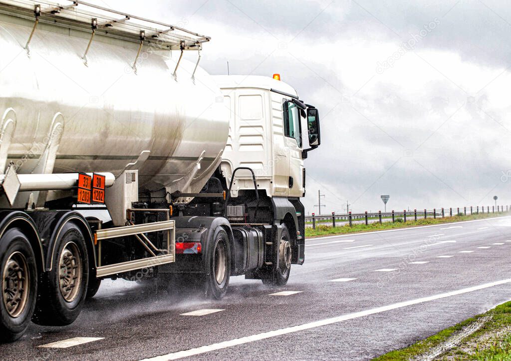 A white semitrailer tractor with a stainless tanker truck transports dangerous goods on the road. Kotsnet transportation of engine oil and diesel fuel to filling stations. Wet road and cloudy rainy day, industry