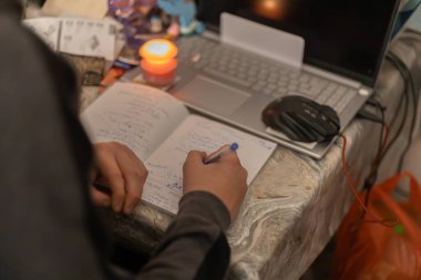 Bir çocuk ödevini mum ışığında çalışmayan bir dizüstü bilgisayarın yanında yapar. Ukrayna 'daki savaş nedeniyle elektrik kesintileri.