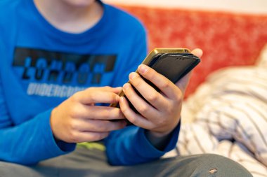 Evde bir yatakta otururken, bir cüzdanda akıllı telefon kullanan bir çocuğun yakın çekimi.