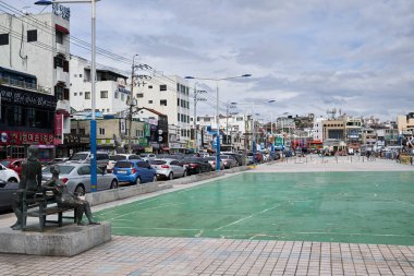 Tongyeong-si, Güney Kore - 25 Eylül 2018: Tongyeong, Güney Kore 'deki Gangguan Limanı' nda, bulutlu bir öğleden sonra gökyüzü altında park edilmiş araçlar ve yerel mağazaların bulunduğu bronz bir heykel yer alıyor..