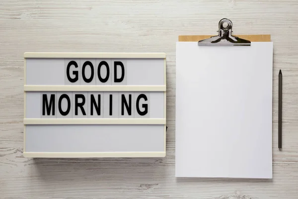 stock image 'Good morning' on a lightbox, clipboard with blank sheet of paper on a white wooden background, top view. Flat lay, overhead, from above. 