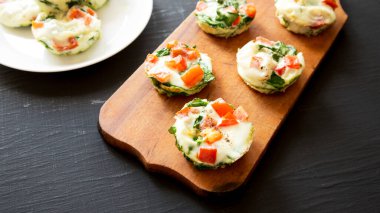 Siyah arka planda, ahşap bir tahtanın üzerinde ıspanaklı ve domatesli ev yapımı yumurta beyazı kahvaltı fincanları..