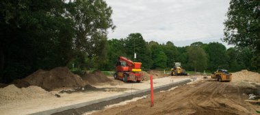 Şehirdeki yeni yol ve kaldırım inşaatı. Sarı traktör ve yol silindiri. Banliyöde yeşil ağaçlar.