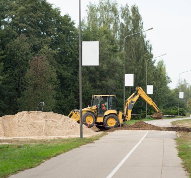 Dünyayı yerinden oynatan ağır ekipmanlar. Sarı kazıcı yol çalışması yapıyor. Bisiklet ve yaya yolu asfalt yolu.