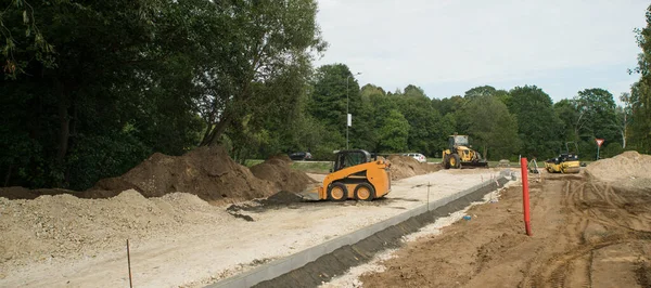Şehirde yeni bir yol inşaatı. Sarı traktör ve yol silindiri. Patinaj yükleyicisi kum yığınlarının yanında. Banliyöde yeşil ağaçlar.
