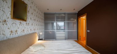 Lüks dairede yatak odasının modern iç mekanı. Rahat yatağın yan manzarası. Büyük sürgülü kapı gardırobu. Komodinin üzerinde lamba.