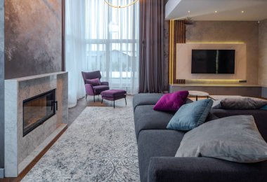 Lüks bir dairede modern bir oturma odası. Şömine ve yastıklı büyük bir kanepe. Duvarda televizyon var. Panoramik pencereler.