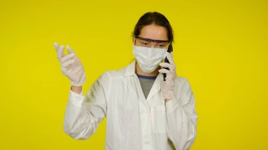 Tıbbi maskeli genç doktor gözlüklü lateks eldivenli hasta ile telefonda konuşuyor.