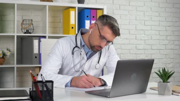 Az orvos fehér orvosi kabátot visel a kórházi irodában, laptopot használ, jegyzetfüzetet ír. — Stock videók
