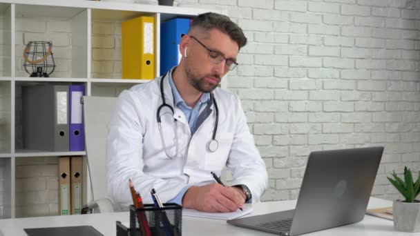 Médico consulta paciente computador portátil chamada de vídeo on-line, escreve sintomas no caderno — Vídeo de Stock