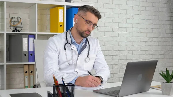 Doctor consults patient online video call laptop, writes symptoms in notebook