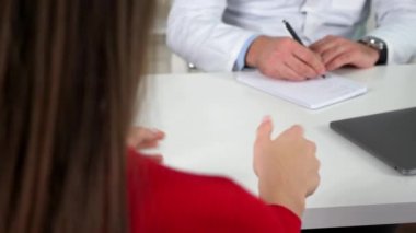 Kadın sağlık sorunlarından bahsediyor, el hareketi yapıyor, doktor belirtiler yazıyor.