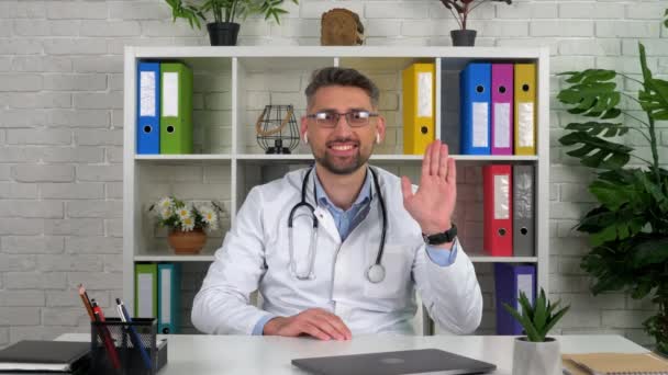 Médico en la oficina del hospital saluda a mano escucha paciente video llamada en línea portátil — Vídeos de Stock