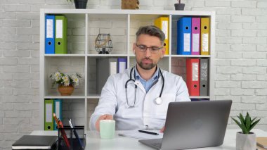 Doktor internetteki hastayı dinliyor. Bilgisayarlı web kamerası, elinde fincan kahveyle.