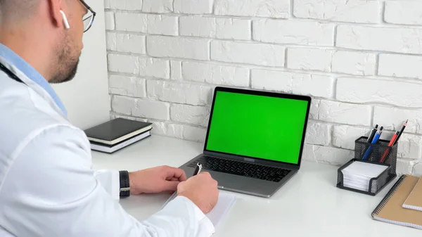 Doctor looking green screen laptop listens patient writes symptoms in notebook