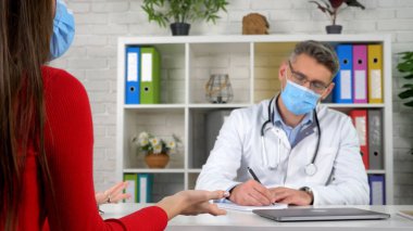 Doktor koruyucu maske takıyor deftere semptomlar yazıyor hastayı dinliyor.