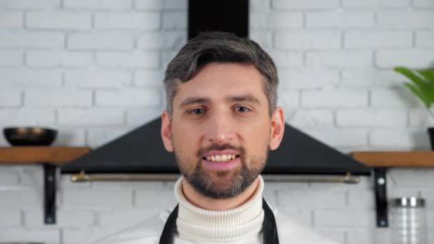 Närbild leende chief man i vit tröja tittar kamera hemma kök — Stockvideo
