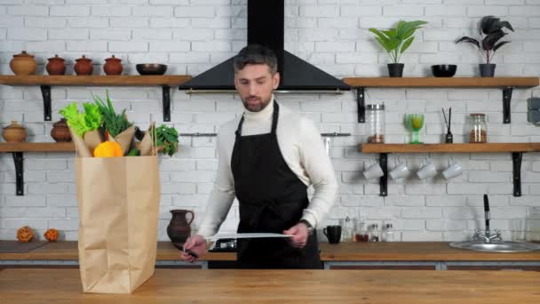 Homem chef em avental preto coloca faca e placa para cortar legumes na cozinha — Vídeo de Stock