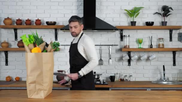 L'uomo in grembiule nero mette coltello e tavola per tagliare verdure sul tavolo della cucina — Video Stock