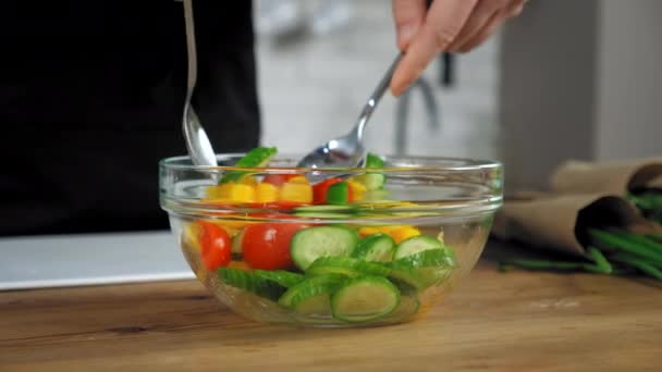 Chef de pé perto da mesa da cozinha misturar salada vegetariana com colheres em tigela de vidro — Vídeo de Stock