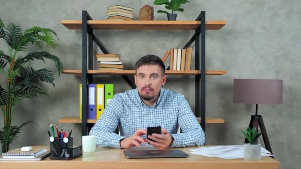 Divertido hombre de negocios sentado en la silla en el escritorio en la oficina sacude la cabeza utiliza smartphone — Vídeos de Stock