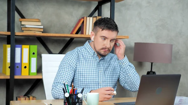 Business man in home office puts on wireless earphones starts using laptop