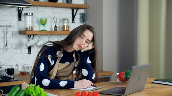 Tired woman sleeps sitting on chair at kitchen table, study online video laptop