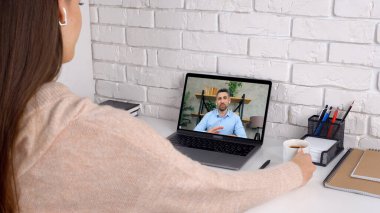 Evde elinde fincan kahveyle bir kadın, uzaktan kumandayla web kamerası görüntülü arama laptopuyla çalışıyor.