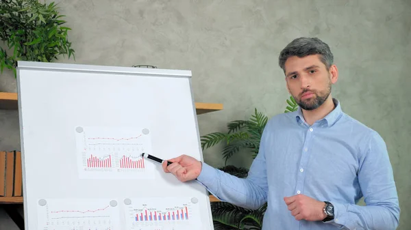 Man teacher points marker diagram on flip chart looking camera teaches videocall