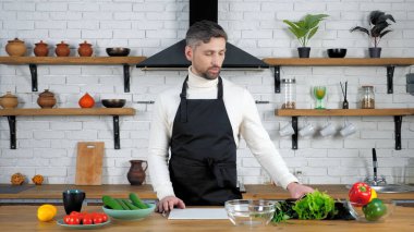 Ev mutfağındaki siyah önlüklü adam cam kaseye marul yaprakları koyar.