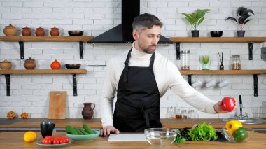 Mutfakta siyah önlüklü adam cam kaseye kırmızı dolma biber koyuyor.