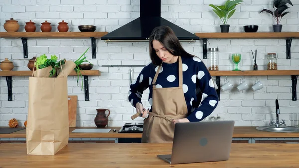 Vrouwelijke chef-kok komt naar huis keuken zet op schort, voor te bereiden online cursus in laptop — Stockfoto