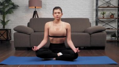 Spor kıyafetli sakin genç bir kadın oturma odasında yoga yapıyor.