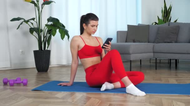 Zooma in sportig kvinna som sitter på yogamattan använder smartphone vänder huvudet och ler — Stockvideo