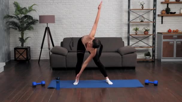 Spor kıyafetli genç bir kadın evdeki yoga minderinde antrenman yapmadan önce ısınıyor. — Stok video