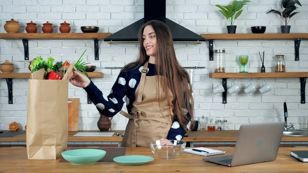 Glimlachende vrouw huisvrouw in beige schort pakt ingrediënten uit papieren zak — Stockfoto