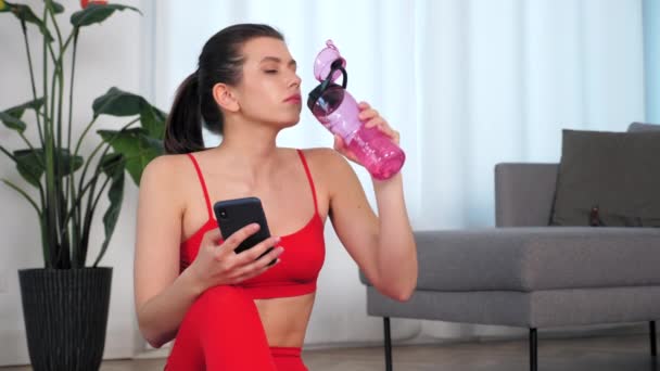 Donna sportiva bere acqua dopo l'esercizio fisico attivo utilizza smartphone a casa — Video Stock