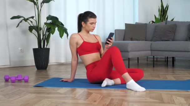 Camera zoom in sportieve serieuze vrouw zitten op yoga blauwe mat maakt gebruik van smartphone — Stockvideo