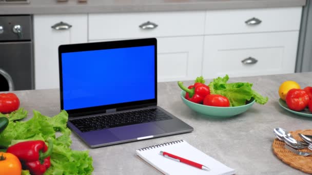 Laptop tela azul fica na mesa da cozinha perto de legumes e notebook com caneta — Vídeo de Stock