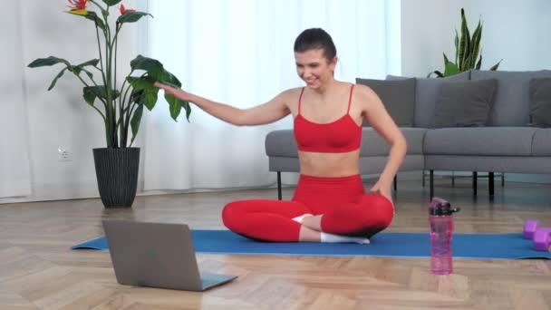 Glimlachende vrouw in sportkleding zittend op yoga mat maakt golf met de handen thuis — Stockvideo