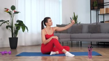Smiling woman sitting on yoga mat greets record video on front camera smartphone