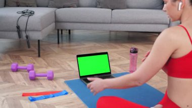 Fit kadın yoga minderinde oturur dizüstü bilgisayara bakar, fitness antrenörüne video çağrısı yapar.