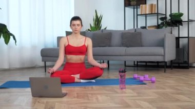 Spor kıyafetli sakin genç bir kadın oturma odasında yoga yapıyor.