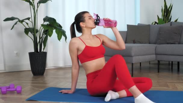 Zoom en mujer deportiva sentada en yoga mat bebidas agua vuelta cabeza mirando cámara — Vídeo de stock
