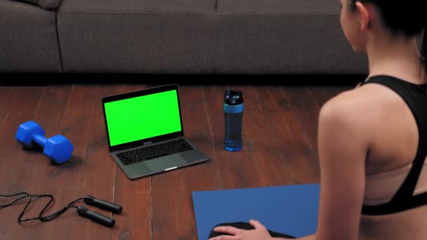 Femme assise sur tapis de yoga regarde écran vert ordinateur portable, salue dit entraîneur de fitness — Video