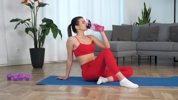 Zoom en mujer deportiva sentada en yoga mat bebidas agua vuelta cabeza mirando cámara — Vídeos de Stock