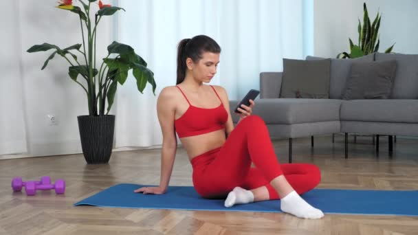 Zoom en mujer deportiva sentada en estera de yoga utiliza smartphone gira la cabeza y sonríe — Vídeo de stock