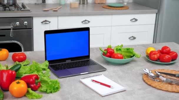 青い画面のラップトップは、ペンで野菜やノートブックの近くのキッチンテーブルの上に立つ — ストック動画