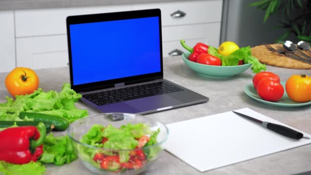 Μπλε οθόνη laptop στο τραπέζι της κουζίνας κοντά σε λαχανικά και ξύλο κοπής με μαχαίρι — Αρχείο Βίντεο
