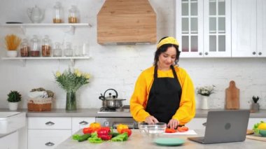 Smiling woman housewife slices tomato listen chef study online video call laptop